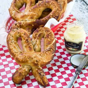 Soft pretzels and a jar of mustard sit on a red checked paper napkin. A basket of pretzels sits in the background. A spreading knife sits next to the jar.