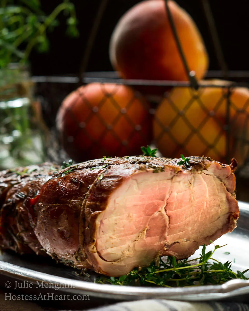 A sliced pork loin with diced peach rum sauce sits on a silver platter showing the light pink center. A basket of peaches sits in the background.