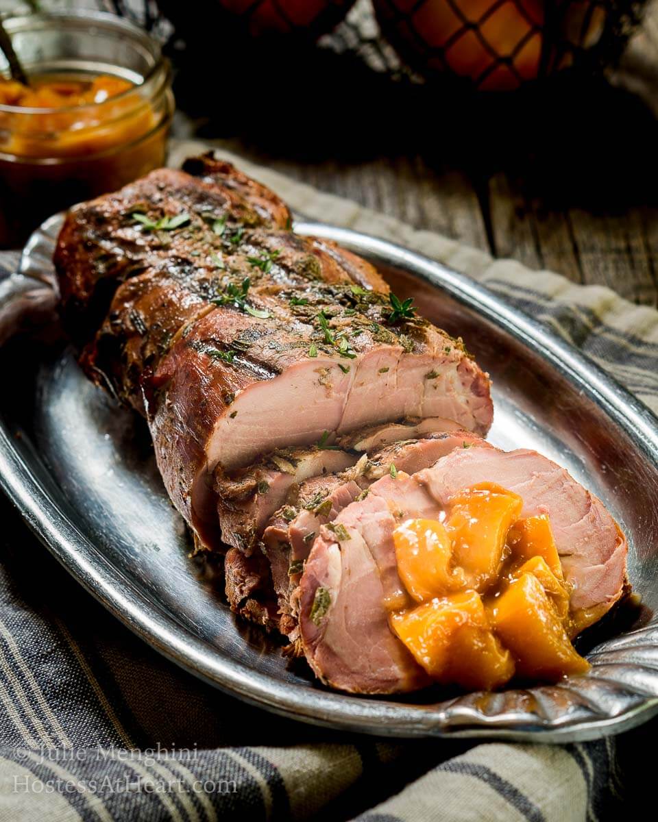 A sliced pork loin with diced peach rum sauce sits on a silver platter. A jar of the rum sauce sits in the background.
