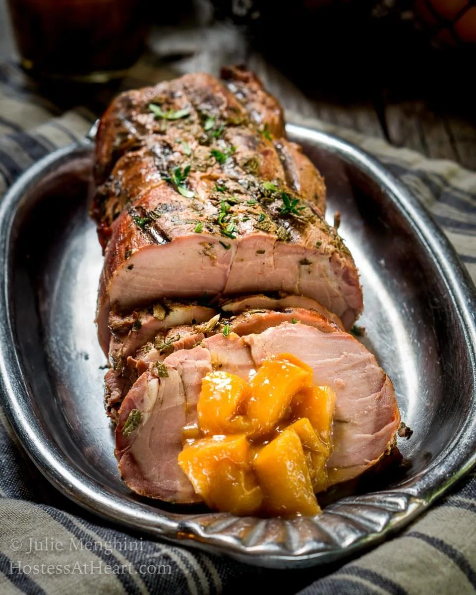 A sliced pork loin with diced peach rum sauce sits on a silver platter over a blue striped towel.