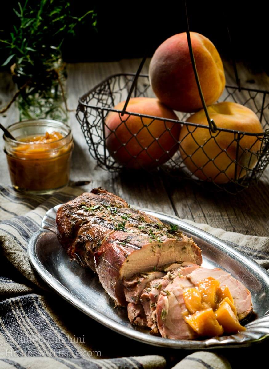 A sliced pork loin with diced peach rum sauce sits on a silver platter. A basket of peaches sits in the background next to a jar of the rum sauce.