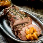 A silver tray holding a Pork Loin with slices leaning against the loin with diced peaches in rum sauce dolloped on the slices.
