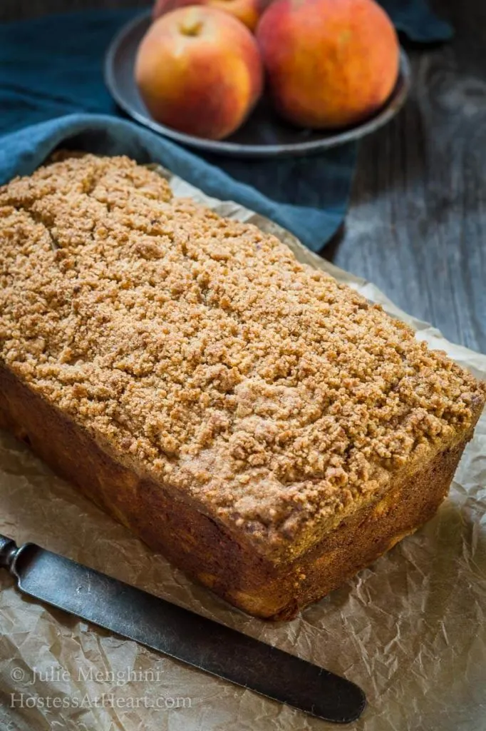 3/4 view of an unsliced loaf of peach bread covered in a crunchy cinnamon-brown sugar streusel. A bowl of peaches sit behind it and an antique knife next to it.