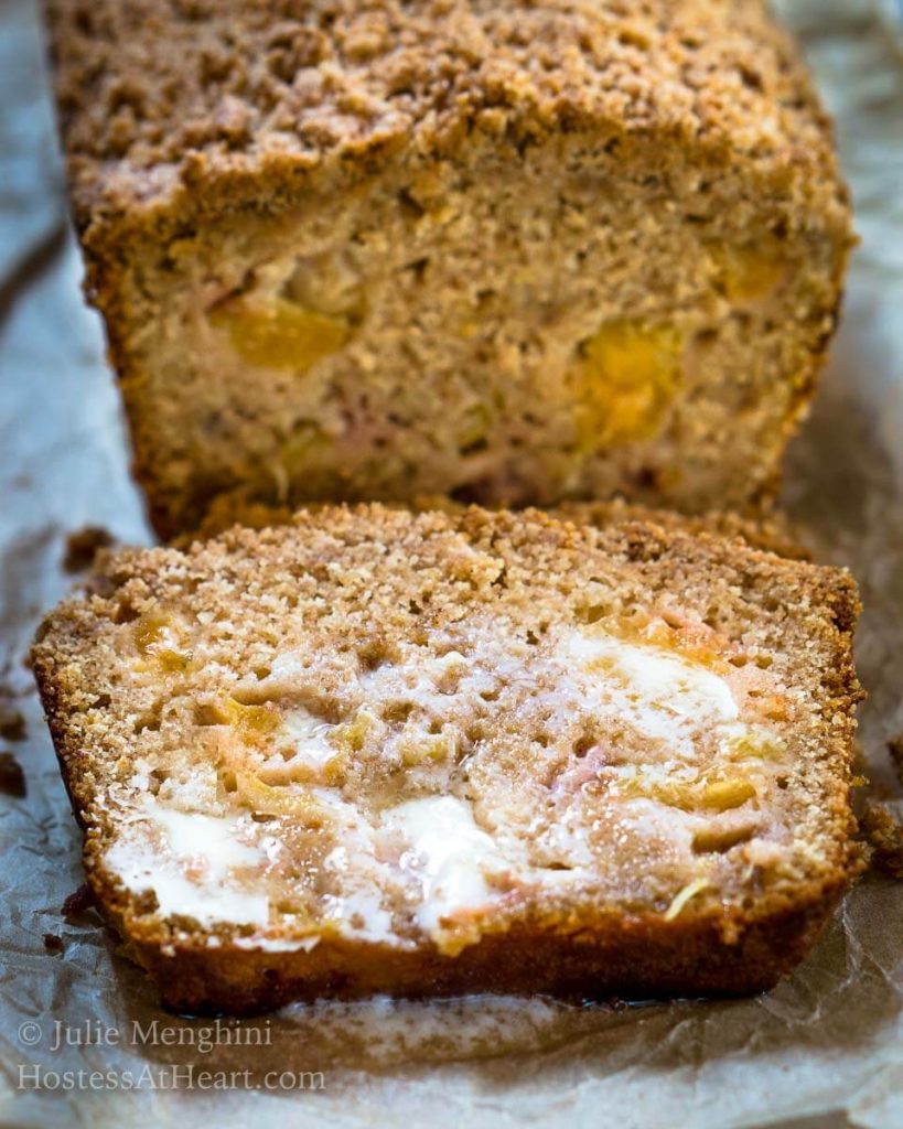 Front angle shot of a slice of peach bread shows cubes of yellow peaches and is covered in melted butter. The bread sits on a metal plate.