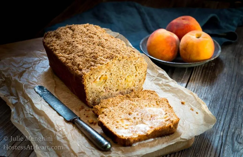 A sliced loaf of streusel bread sitting on a piece of parchment paper with a bowl of fresh peaches next to it.