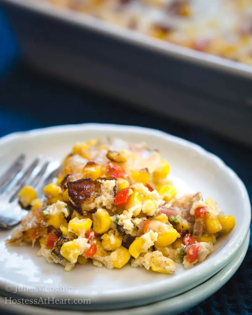 A close up of a plate of Roasted Corn and Bacon Casserole. The casserole dish sits in the background and a fork sits on the plate.