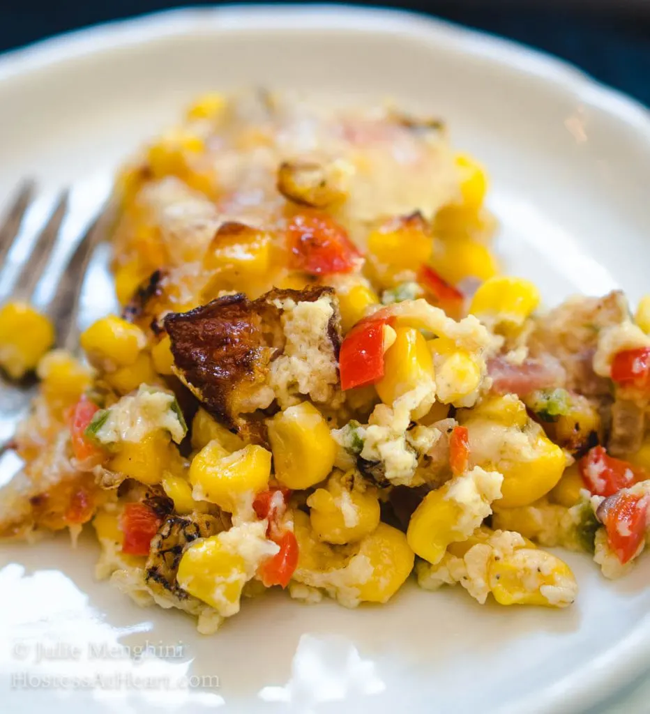 A white plate with a serving of roasted corn and bacon casserole A fork sits next to the food.