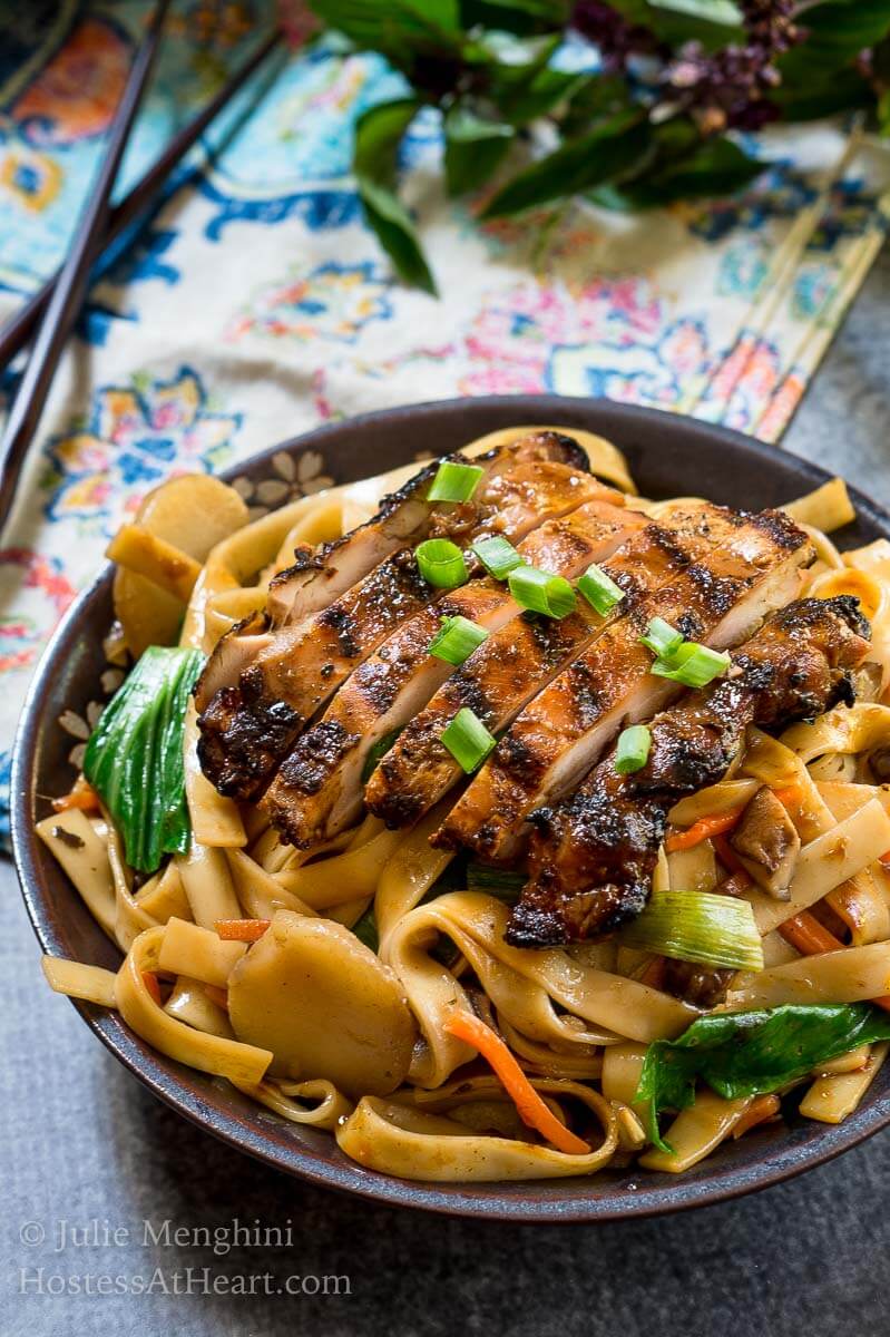 Top side angle view of a dark gray bowl filled with noodles, carrots, and water chestnuts,  sauced in Thai Basil flavors topped with grilled chicken and garnished with sliced green onions. The bowl sits over a multi-colored napkin and a pair of chopsticks. Fresh Thai basil sits in the background.