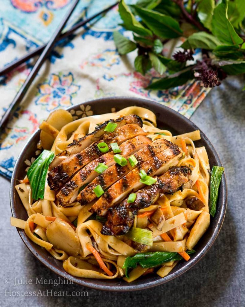 Glistening grilled Chicken sliced over saucy noodles and Asian veggies in a gray bowl. HostessAtHeart.com