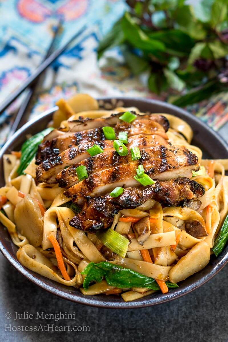 Top angle view of a dark gray bowl filled with noodles, carrots, and water chestnuts,  sauced in Thai Basil flavors topped with grilled chicken and garnished with sliced green onions. The bowl sits over a multi-colored napkin and a pair of chopsticks. Fresh Thai basil sits in the background.