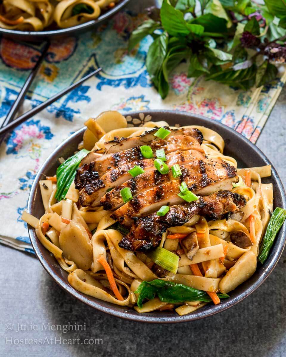 Top angle view of a dark gray bowl filled with noodles, carrots, and water chestnuts,  sauced in Thai Basil flavors topped with grilled chicken and garnished with sliced green onions. The bowl sits over a multi-colored napkin and a pair of chopsticks. Fresh Thai basil sits in the background.