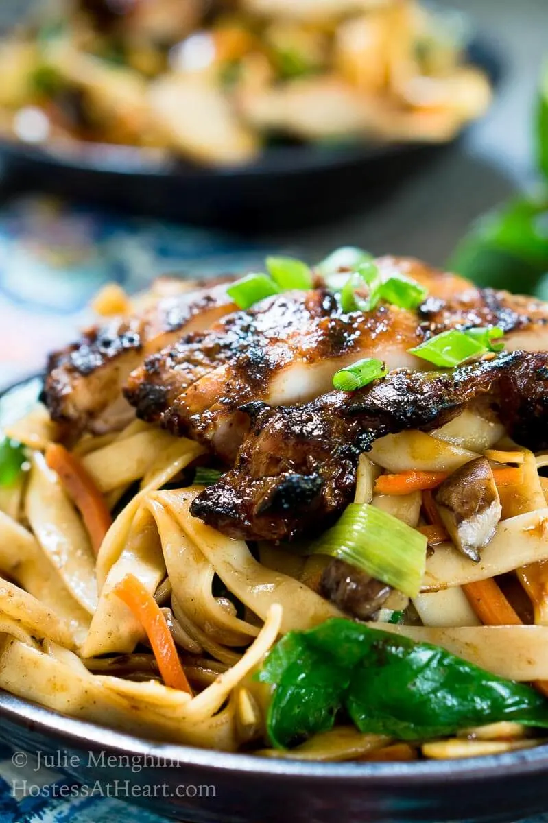 A close up side angle view of a dark gray bowl filled with noodles, carrots, and water chestnuts,  sauced in Thai Basil flavors topped with grilled chicken and garnished with sliced green onions. The bowl sits over a multi-colored napkin and a pair of chopsticks. A second bowl sits in the background.