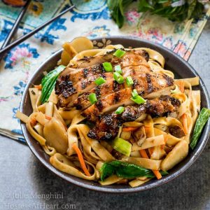 Side angle view of a dark gray bowl filled with noodles, carrots, and water chestnuts,  sauced in Thai Basil flavors topped with grilled chicken and garnished with sliced green onions.