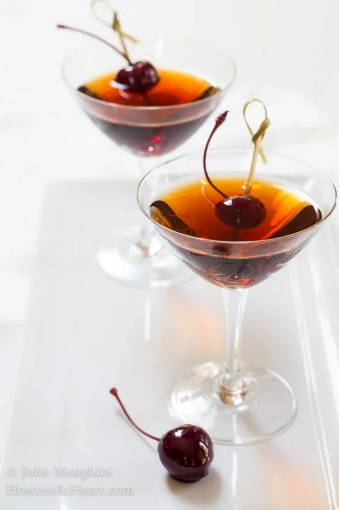 Side view of two martini glasses holding Cuban Manhattans. A Bada cherry sits next to the glass as well as one on a skewer in the drink.
