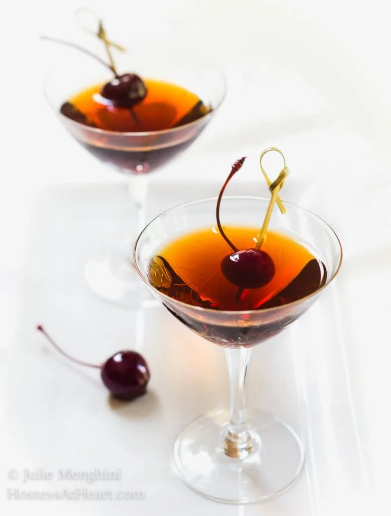 Top-down view of two martini glasses holding Cuban Manhattans. A Bada cherry sits next to the glass as well as one on a skewer in the drink.