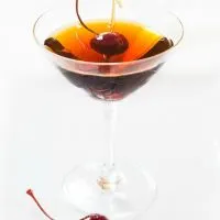 Side view of a martini glass holding a Cuban Manhattan. A Bada cherry sits next to the glass as well as one in the drink.