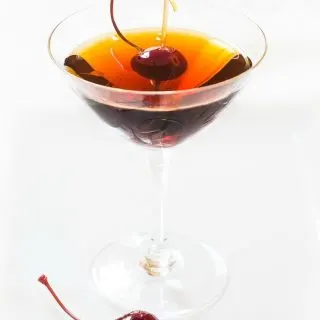 Side view of a martini glass holding a Cuban Manhattan. A Bada cherry sits next to the glass as well as one in the drink.