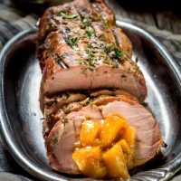 A sliced pork loin with diced peach rum sauce sits on a silver platter over a blue striped towel.