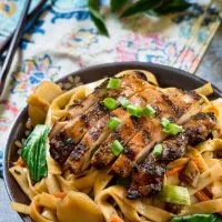 Top side angle view of a dark gray bowl filled with noodles, carrots, and water chestnuts, sauced in Thai Basil flavors topped with grilled chicken and garnished with sliced green onions. The bowl sits over a multi-colored napkin and a pair of chopsticks. Fresh Thai basil sits in the background.