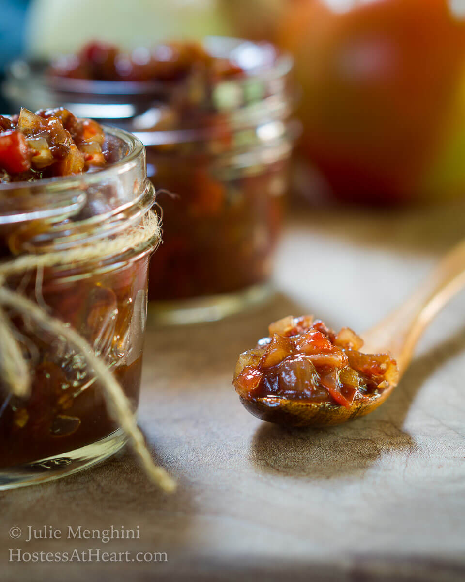 Close up side view of a wooden spoon filled with tomato onion chutney on a wooden board next to 2 jars filled with tomato chutney.