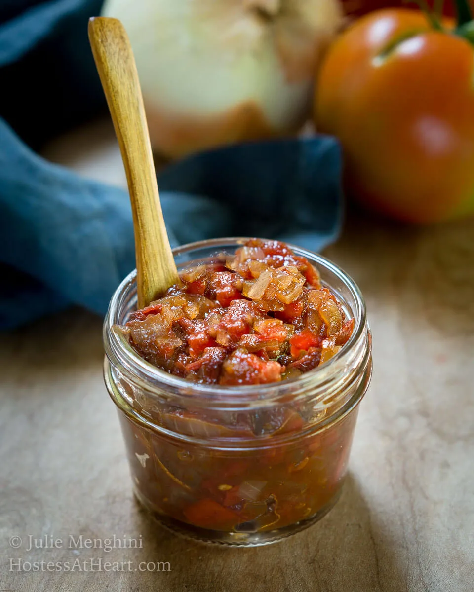 Jar of diced tomatoes and onions in a small jar with a wooden spoon in it. A blue napkin, tomato and onion sits behind it.