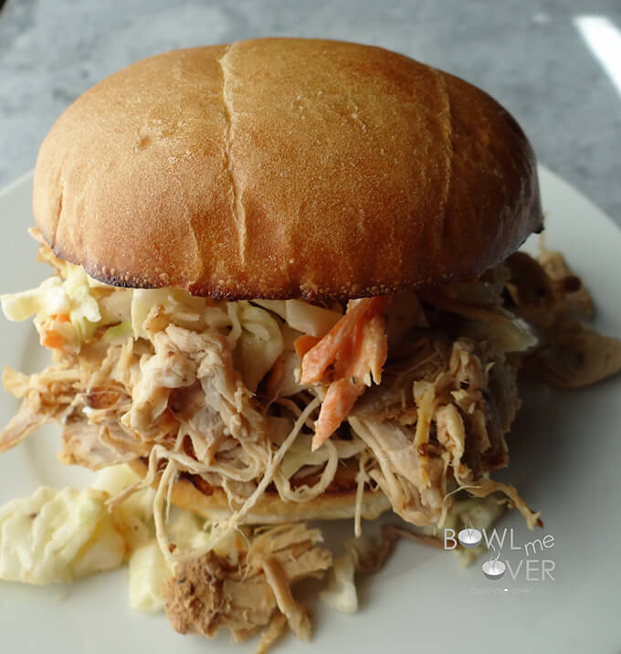 A close-up side view of a Kauai Pulled pork sandwich on a white plate, with Pulled pork spilling out of the bun.