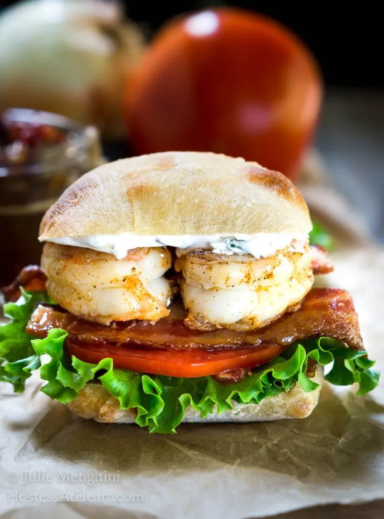 A closeup side view of a sandwich layered with spicy grilled shrimp, bacon, lettuce, tomato, and aioli inside a ciabatta roll. A jar of tomato relish sits in the background.