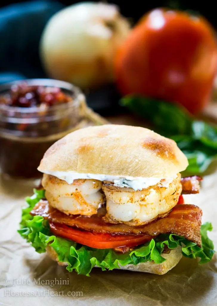 A closeup side view of a sandwich layered with spicy grilled shrimp, bacon, lettuce, tomato, and aioli inside a ciabatta roll. A jar of tomato relish sits in the background.