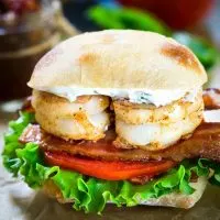 Front view of a sandwich filled with two grilled shrimp over bacon, tomato, and lettuce. The sandwich has a layer of aoili and the bread is a sliced Ciabatta roll.
