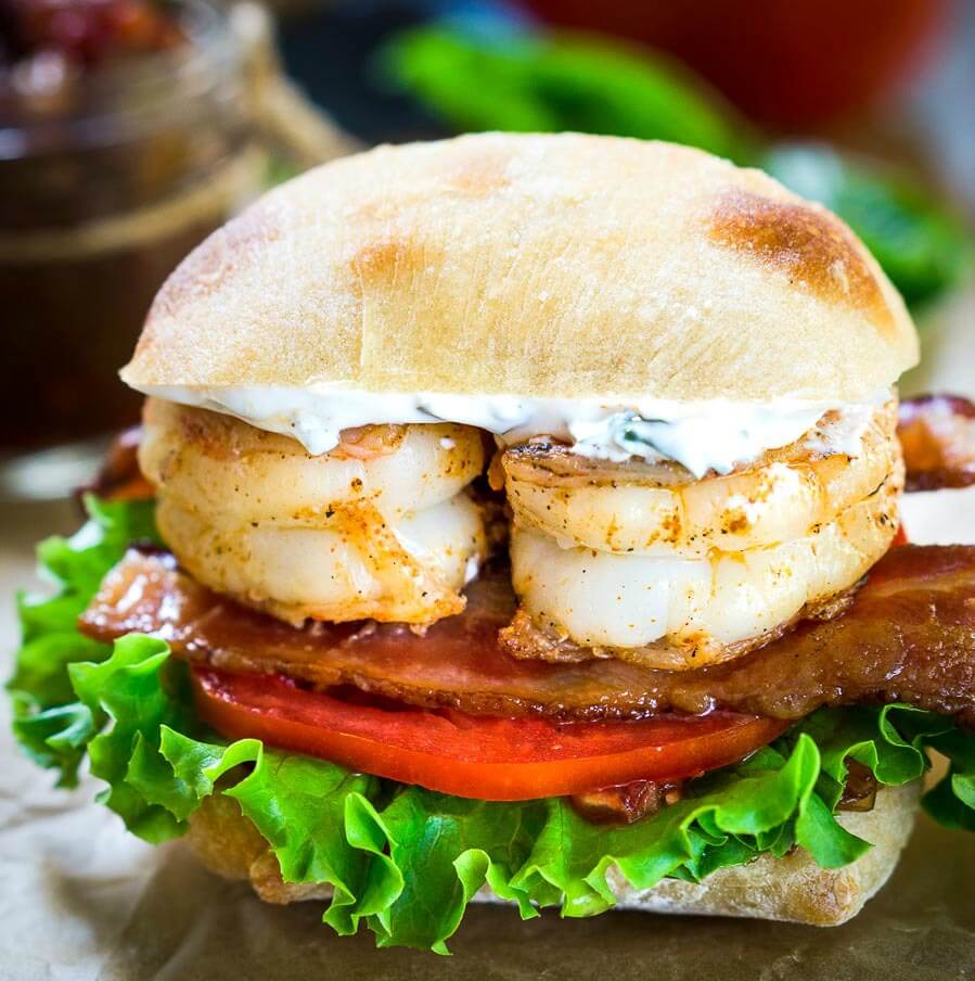 Front view of a sandwich filled with two grilled shrimp over bacon, tomato, and lettuce. The sandwich has a layer of aoili and the bread is a sliced Ciabatta roll.