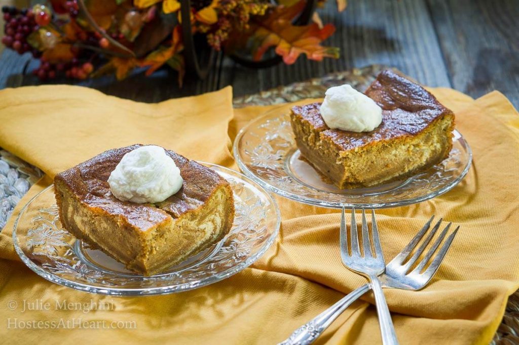 Two pieces of Pumpkin cheesecake with swirls of cream cheese filling running through it and topped with whipped cream sits on glass plates over a gold napkin. Forks sit in the foreground and a fall centerpiece sit in the background.