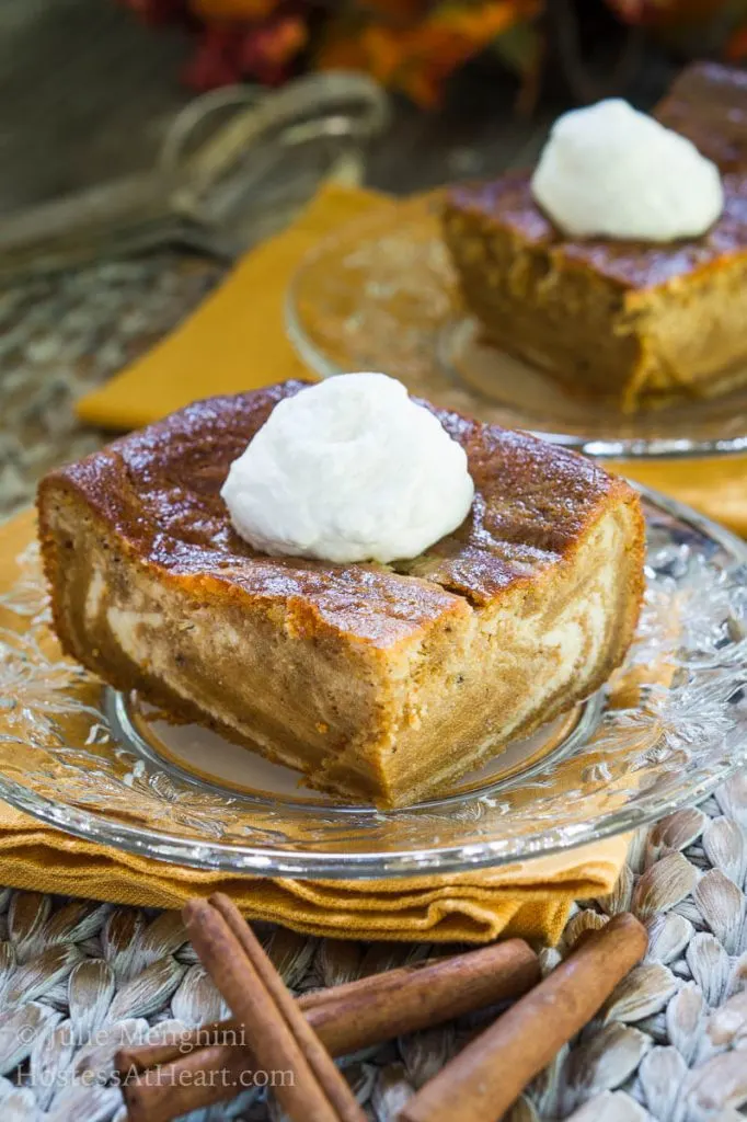 A piece of Pumpkin cheesecake with swirls of cream cheese filling running through it and topped with whipped cream sits on a glass plate over a gold napkin. A second slice sits in the background.