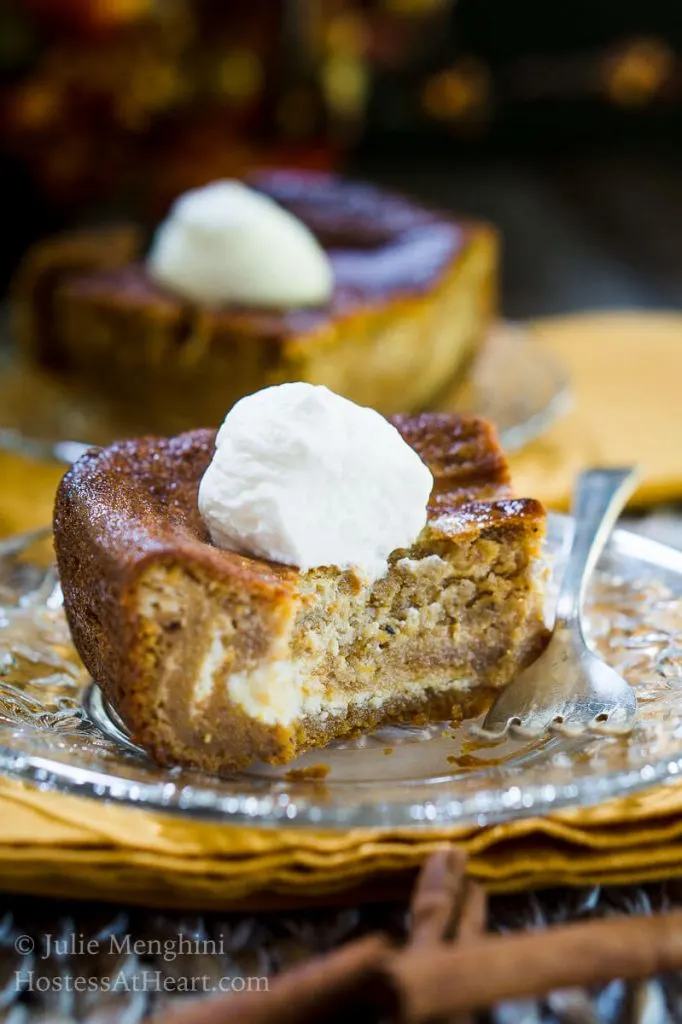 A piece of Pumpkin cheesecake with swirls of cream cheese filling running through it and topped with whipped cream sits on a glass plate over a gold napkin. A second slice sits in the background.