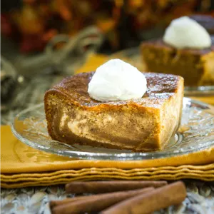A piece of Pumpkin cheesecake with swirls of cream cheese filling running through it and topped with whipped cream sits on a glass plate over a glass plate on a gold napkin. Cinnamon sticks sit in the foreground and a second slice sit in the background.