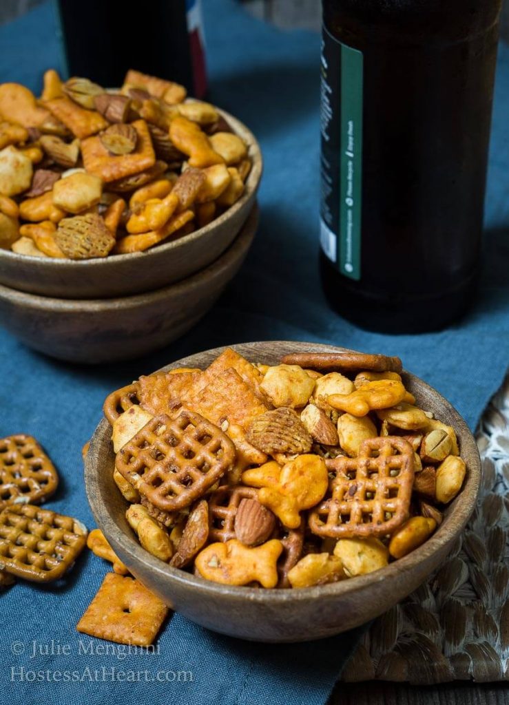 Two wooden bowls filled with Spicy Sriracha Snack Mix over a blue napkin. Additional snack mix is spilled next to the bowls.