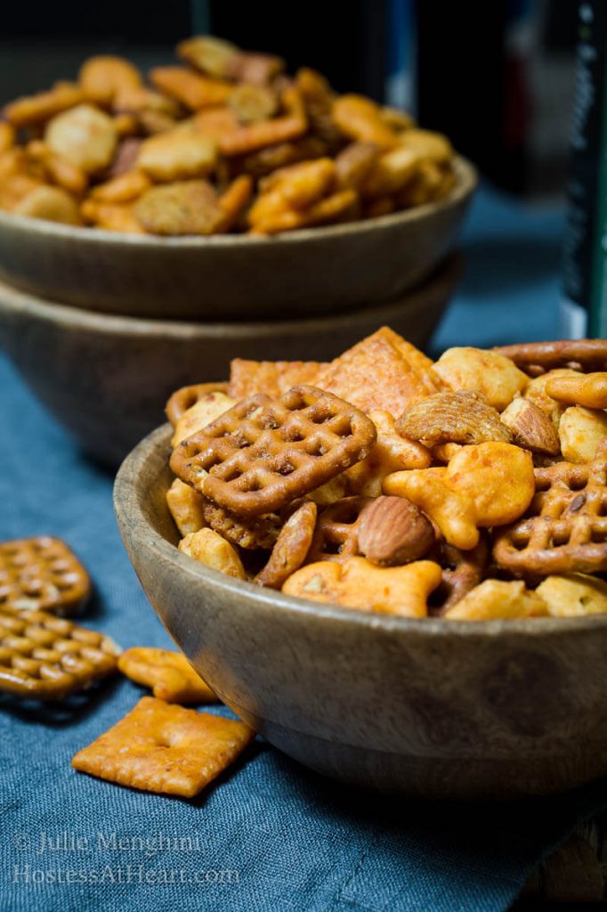 Sideview of two wooden bowls brimming with Chex mix over a blue napkin. Additional Chex mix is spilled next to the bowls.