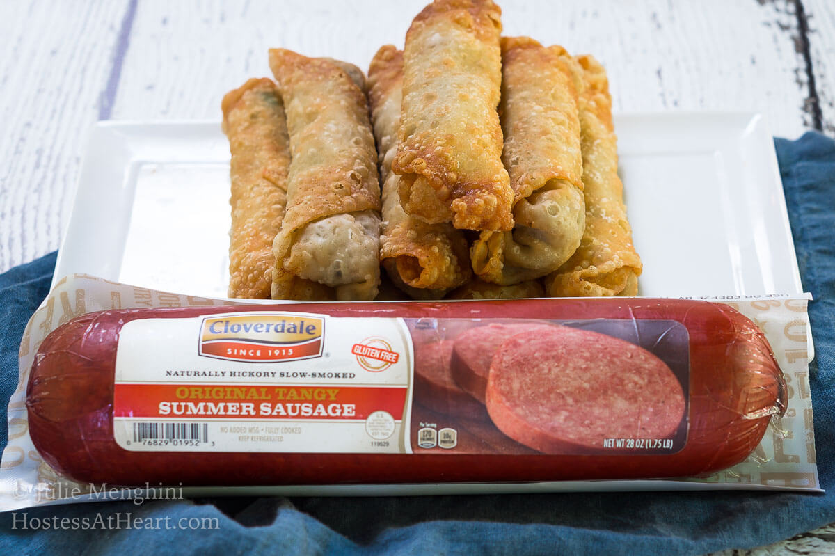A stack of egg rolls sitting behind a roll of Cloverland Summer Sausage over a blue napkin.