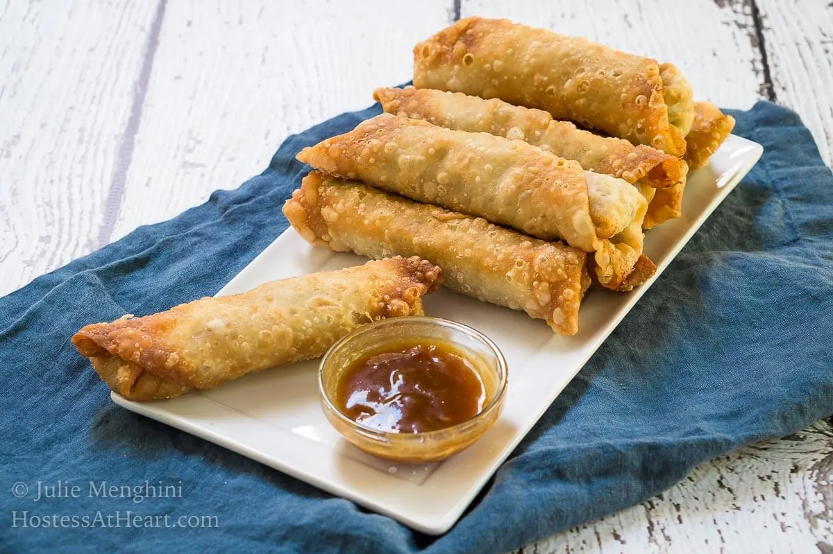 A stack of egg rolls and a small dish of dipping sauce sitting on a white plate over a blue napkin.