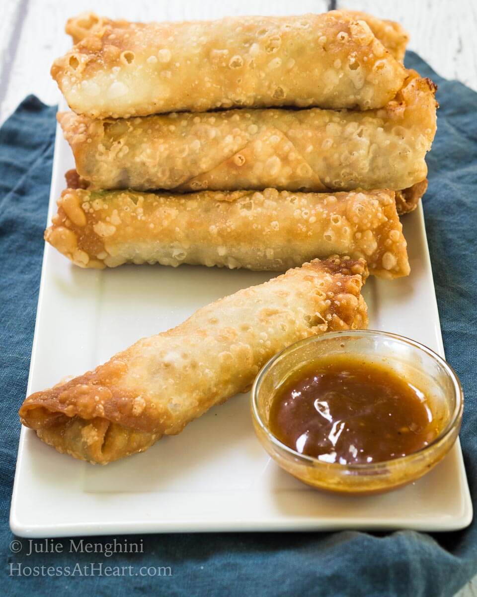 A stack of egg rolls and a small dish of dipping sauce sitting on a white plate over a blue napkin.