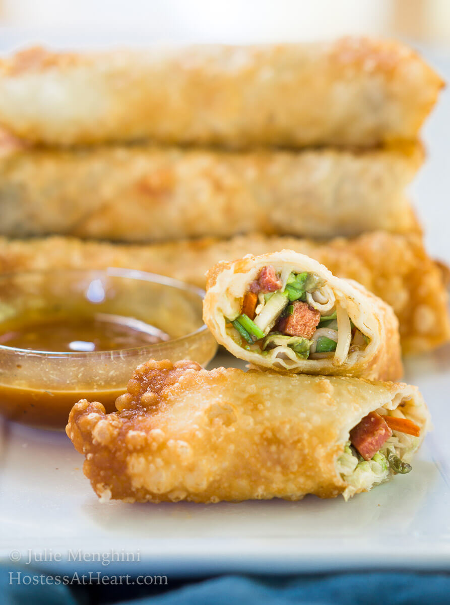 An egg roll cut in half showing the filling which includes sausage and bean sprouts next to a bowl of dipping sauce. A stack of egg rolls sits in the background.