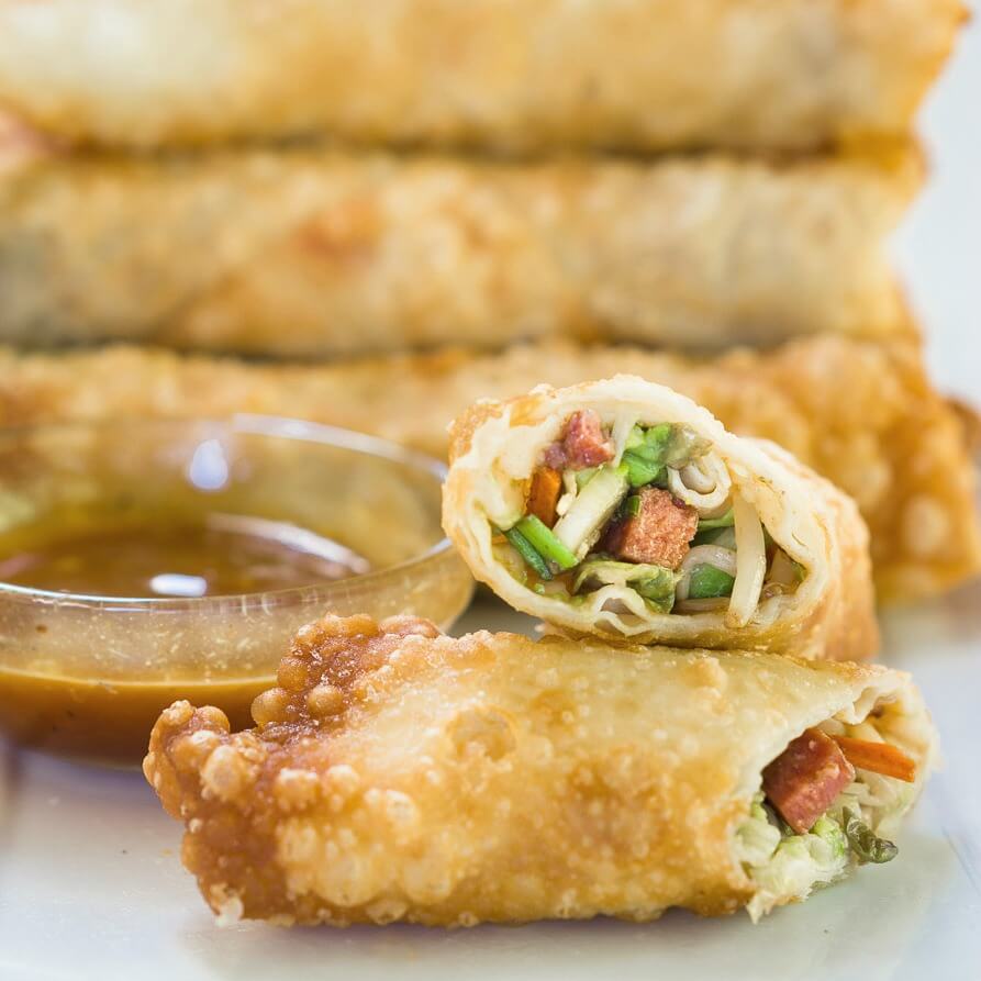 An egg roll cut in half showing the filling which includes sausage and bean sprouts next to a bowl of dipping sauce. A stack of egg rolls sits in the background.