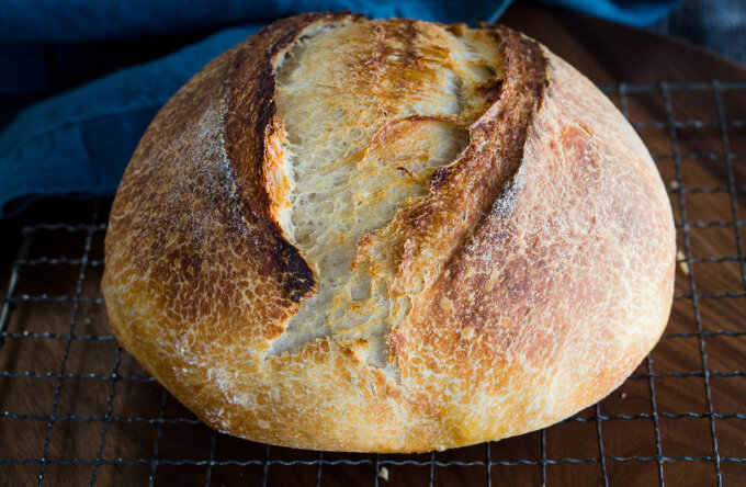 Sideview of a round loaf of baked and browned sourdough bread sitting on a cooling rack over a blue napkin.