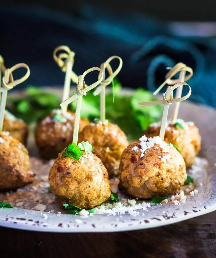 A table view of 6 meatballs with wooden skewers inserted through the top of each meatball sitting on a gray plate garnished with parsley and grated cheese in front of a blue napkin.