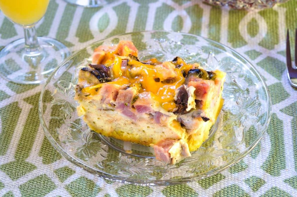 Top angled view of a piece of Ham and Cheese strata with melted cheese and mushrooms sitting on a clear glass plate over a green and white placemat. 