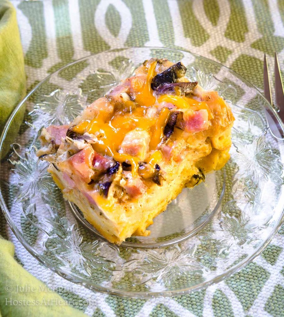 Top-down view of a piece of Ham and Cheese strata with melted cheese and mushrooms sitting on a clear glass plate over a green and white placemat. 