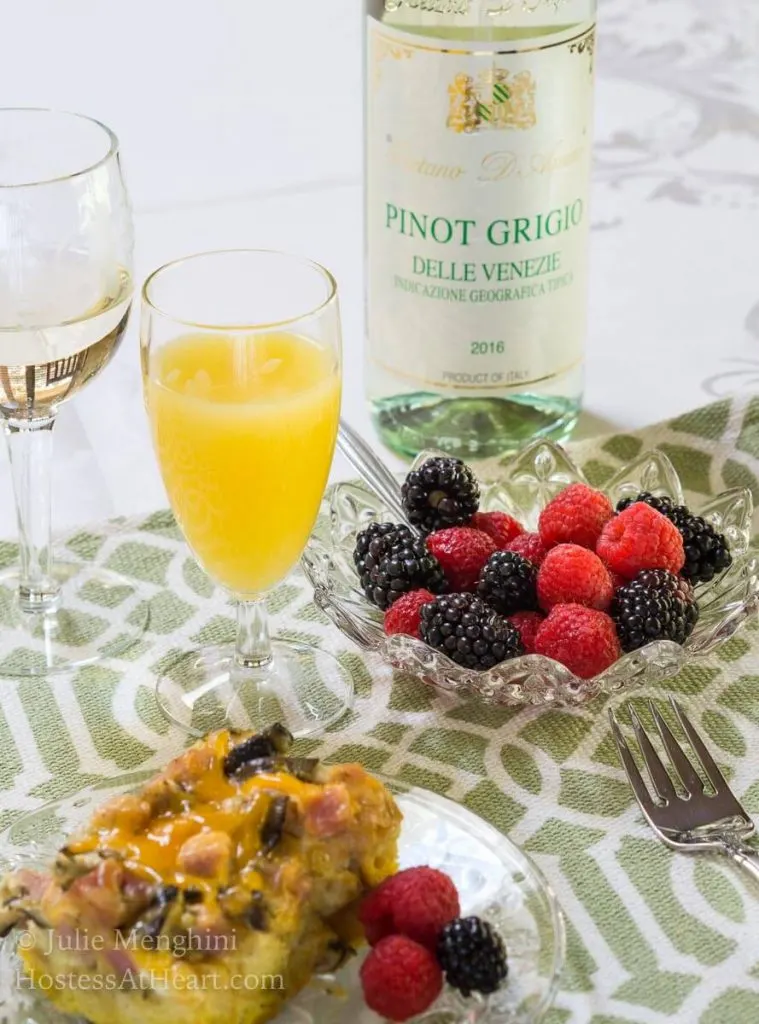 Top angled view of a piece of Ham and Cheese strata with melted cheese and mushrooms sitting on a clear glass plate over a green and white placemat. Fresh raspberries and blackberries sit next to it on the plate and in a bowl behind it with a glass of mimosa and a bottle of white wine.