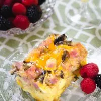 Top angled view of a piece of Ham and Cheese strata with melted cheese and mushrooms sitting on a clear glass plate over a green and white placemat. Fresh raspberries and blackberries sit next to it on the plate and a bowl of the berries sit in the background.