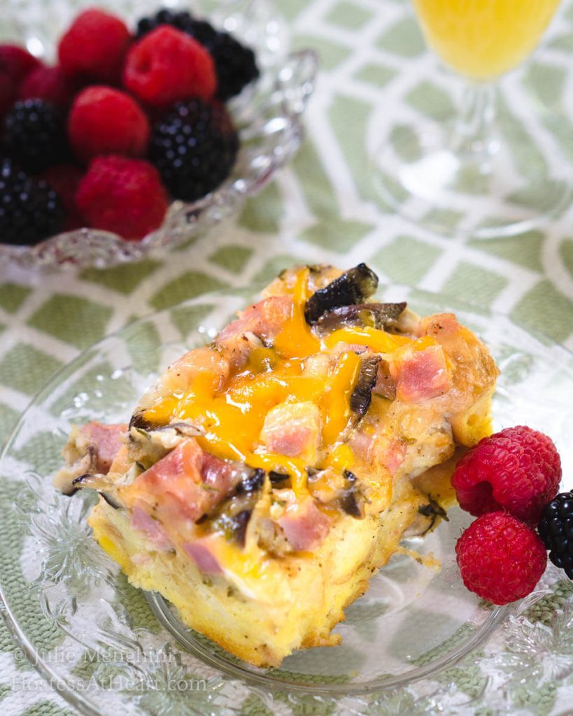 Top angled view of a piece of Ham and Cheese strata with melted cheese and mushrooms sitting on a clear glass plate over a green and white placemat. Fresh raspberries and blackberries sit next to it on the plate and in a bowl behind it.