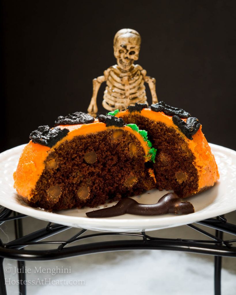 A chocolate cake filled with peanut butter chips on a white plate that's decorated as a pumpkin face. A skeleton sits behind it.