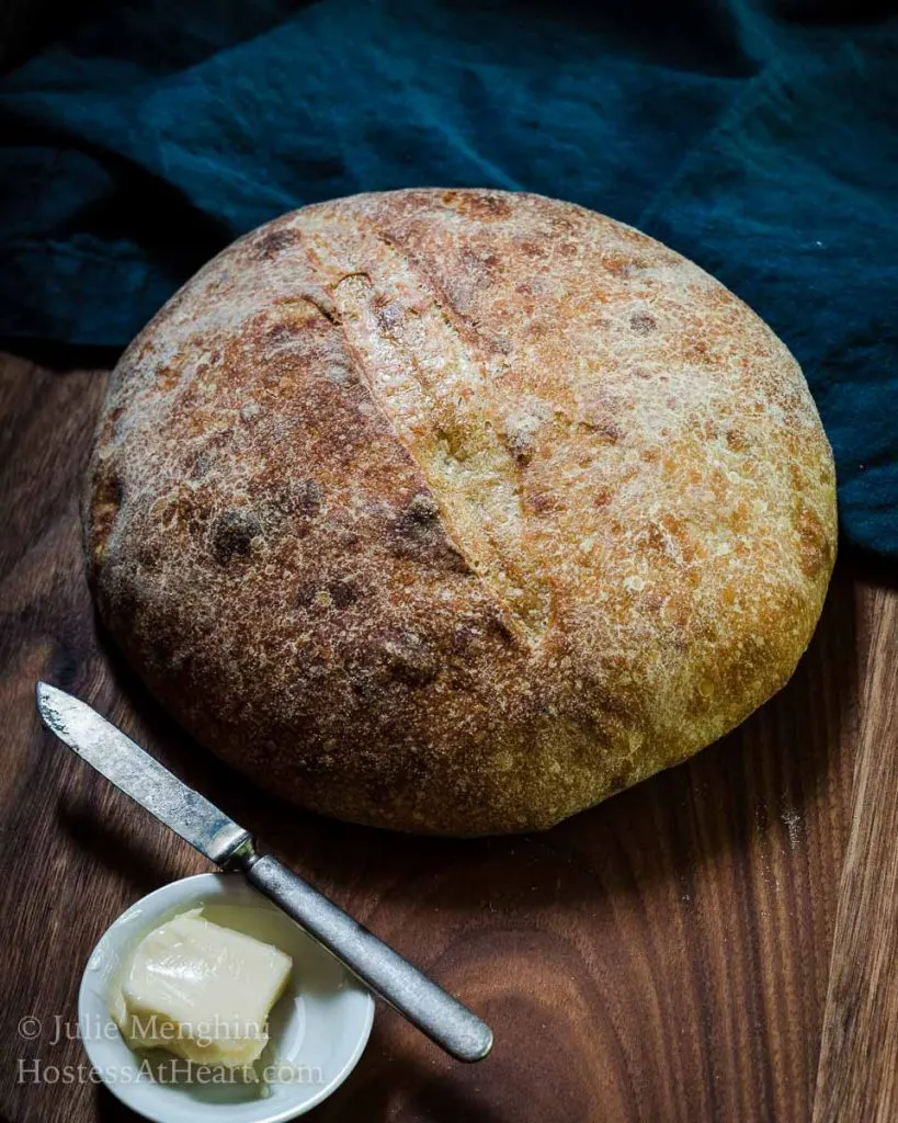 Front view of a round loaf of sourdough showing a browned crispy crust sitting on a cooling rack. A pad of butter sits in a white dish with a butter knife off to the side. The title runs across the bottom.